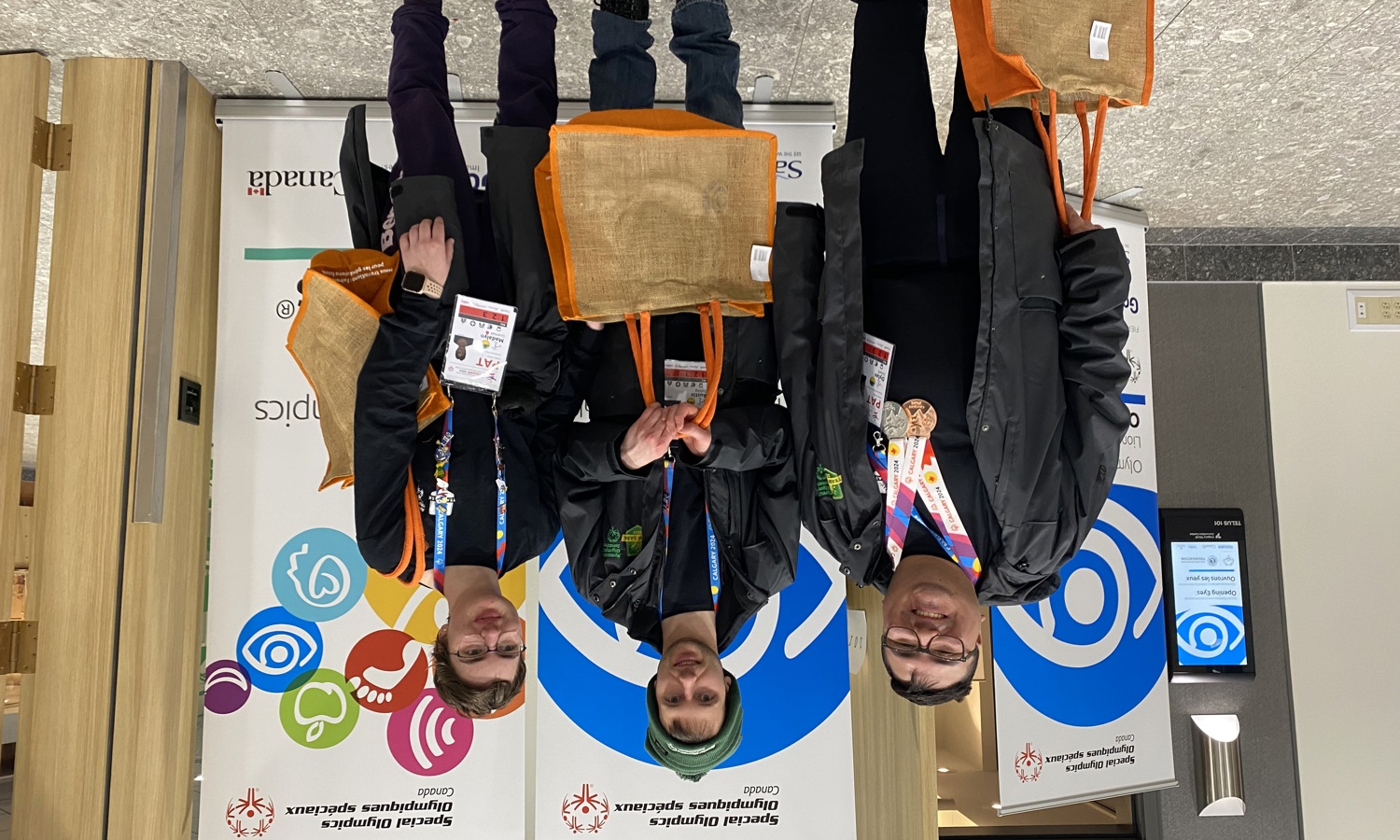Three athletes are standing in front of Healthy Athletes banners and smiling for the camera. They are holding orange tote bags, and wearing all black with colourful lanyards around their necks.