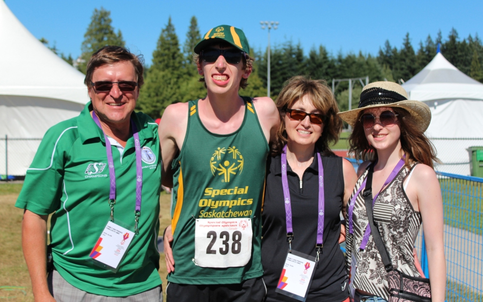 A family of four, posing for a group photo. From left to right is dad, the athlete, mom, and sister. The athlete is wearing a Special Olympics Saskatchewan tank top with the number 238 pinned to his shirt. His family members are wearing colourful lanyards to show that they are with him as supporters.