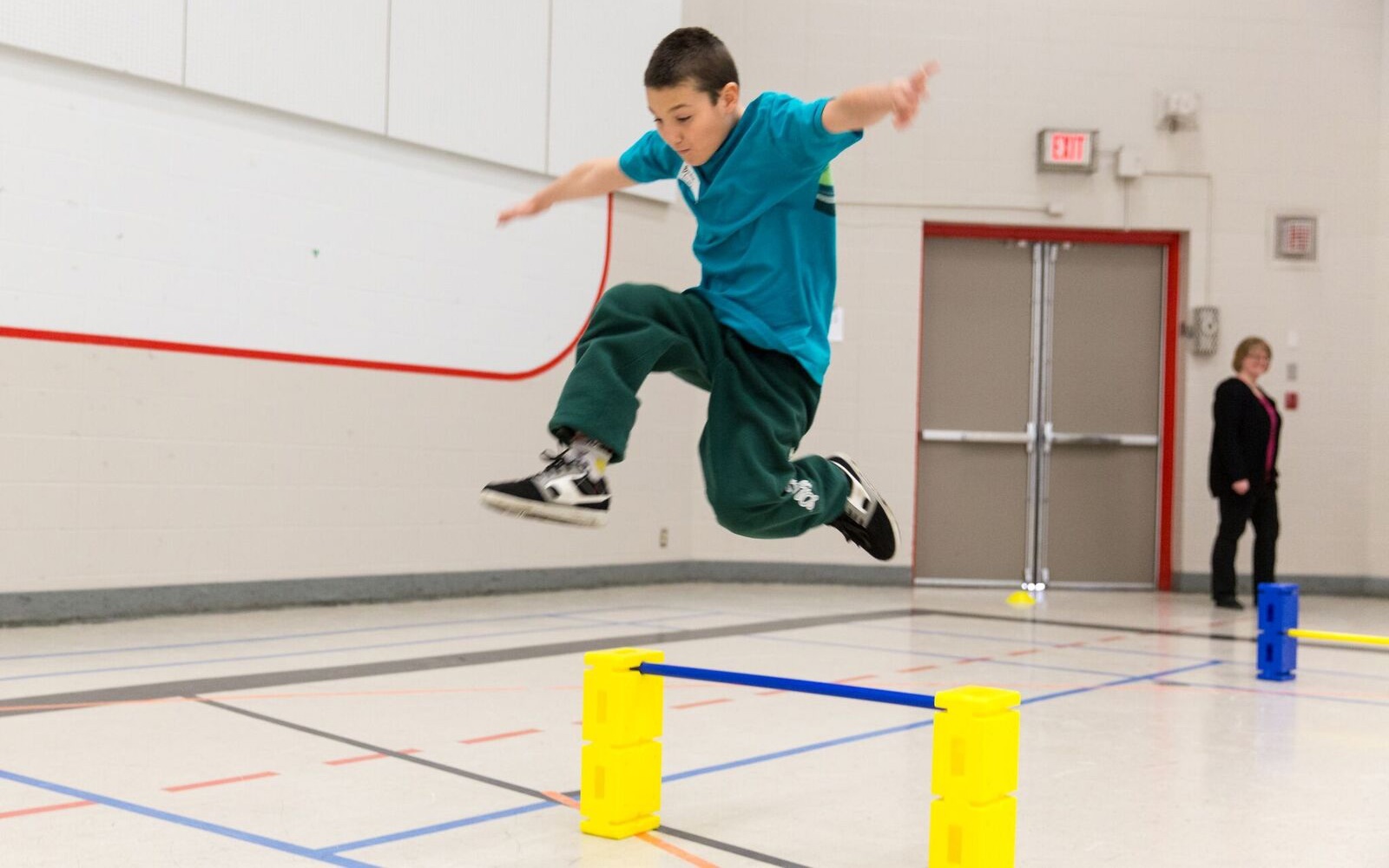 A young athlete in a teal blue t-shirt is jumping over a hurdle in a gymnasium.