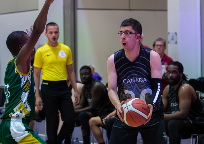 An action shot photo of a basketball game. An athlete in a "Canada" basketball jersey has possession of the basketball. An athlete on the opposing team is trying to defend their team. In the background you can see a referee with a whistle in his mouth as well as some other players on the bench.