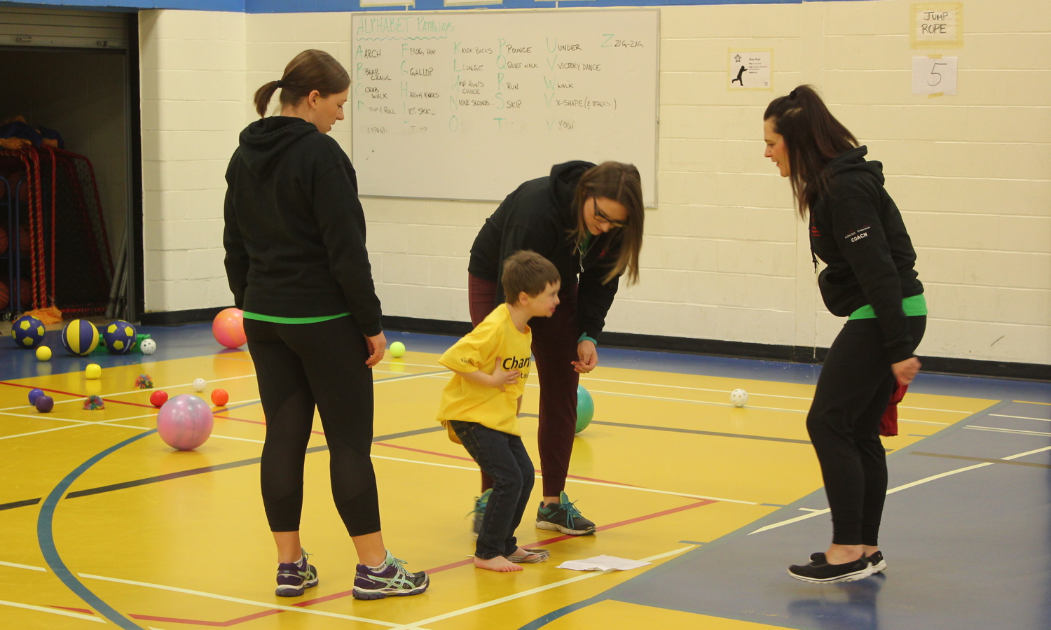 Three volunteers and coaches are standing around a young athlete, assisting him with the activity. They are all standing in a gymnasium and a variety of different sized and coloured balls can be seen in the background.