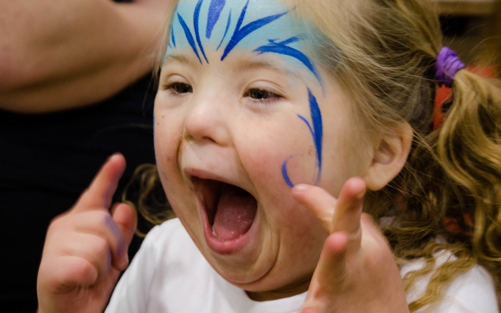 A young girl athlete has her face painted with blue paint on her forehead and cheeks, shows a very happy expression. She is turned diagonally to the left away from the camera, but you can still see her face. She is wearing a white Elsa t-shirt.