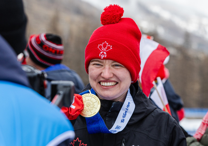 A close up image of an athlete smiling for a camera that is out of focus of the image. She is holding up the gold medal that is hanging around her neck. She is also wearing a red toque and mittens, as well as a black winter jacket.