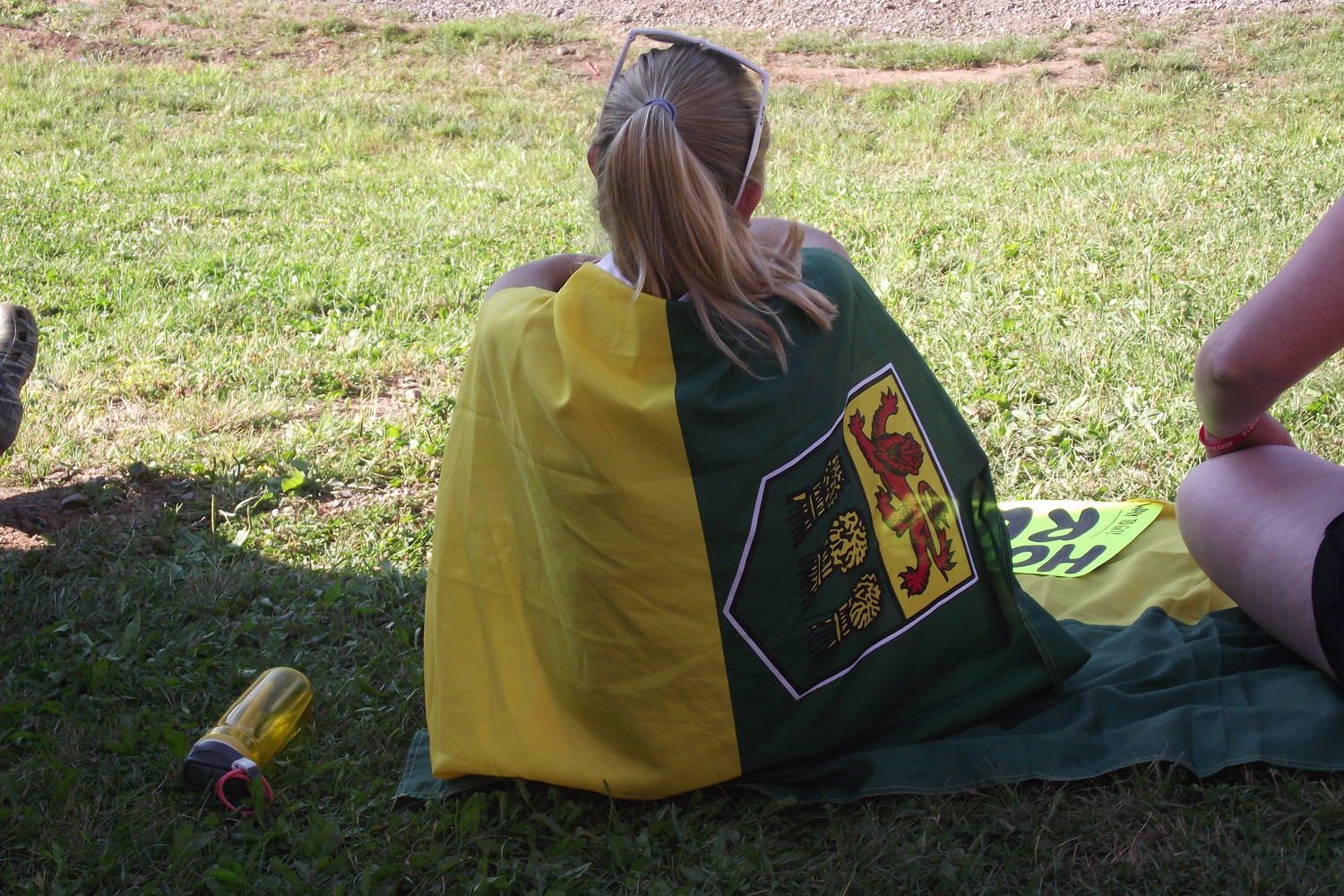 A girl is sitting on the grass with her back towards the camera. Draped around her like a cape is the Saskatchewan flag.