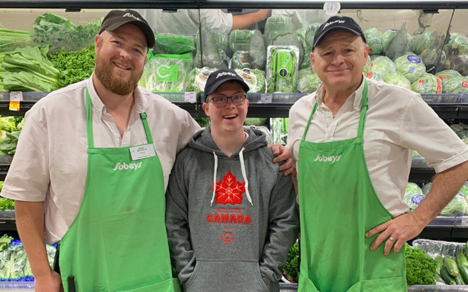 Three men standing beside each other. The two men on either end are wearing white shirts and green aprons with the Sobeys logo. The man in the middle is wearing a gret hoodie with Canada logo. All are smiling at the camera. 
