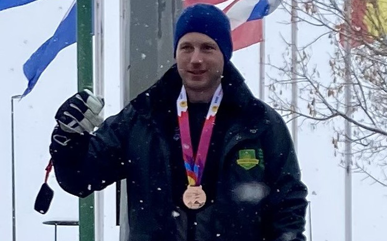 A photo of Taylor, a Special Olympics Saskatchewan athlete. He is standing outside wearing a winter jacket and hat. There is snow falling around him and you can see flags waving behind him. He has a medal around his neck and putting his arm up.