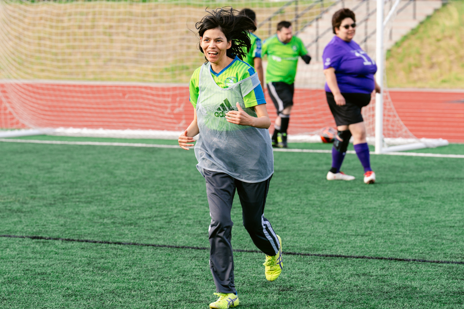 A female soccer player happily running on the soccer field. She is wearing a bright green jersey with a clear grey pinnie over top of it.  She is also wearing bright yellow soccer cleats. Behind her is the soccer net and three other soccer players out of focus.