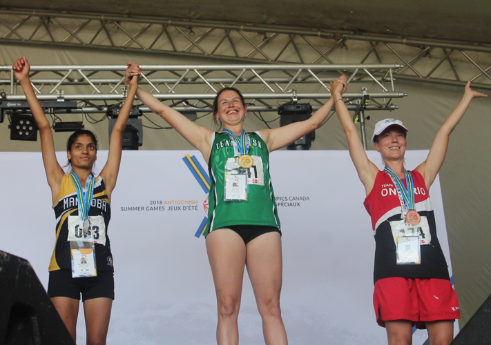 Three Special Olympics athletes standing on the podium. Saskatchewan's athlete in the middle has won gold. Manitoba's athlete on the left has won silver. Ontario's athlete on the right has won bronze. All three athletes are holding hands and holding them up in the winners position.