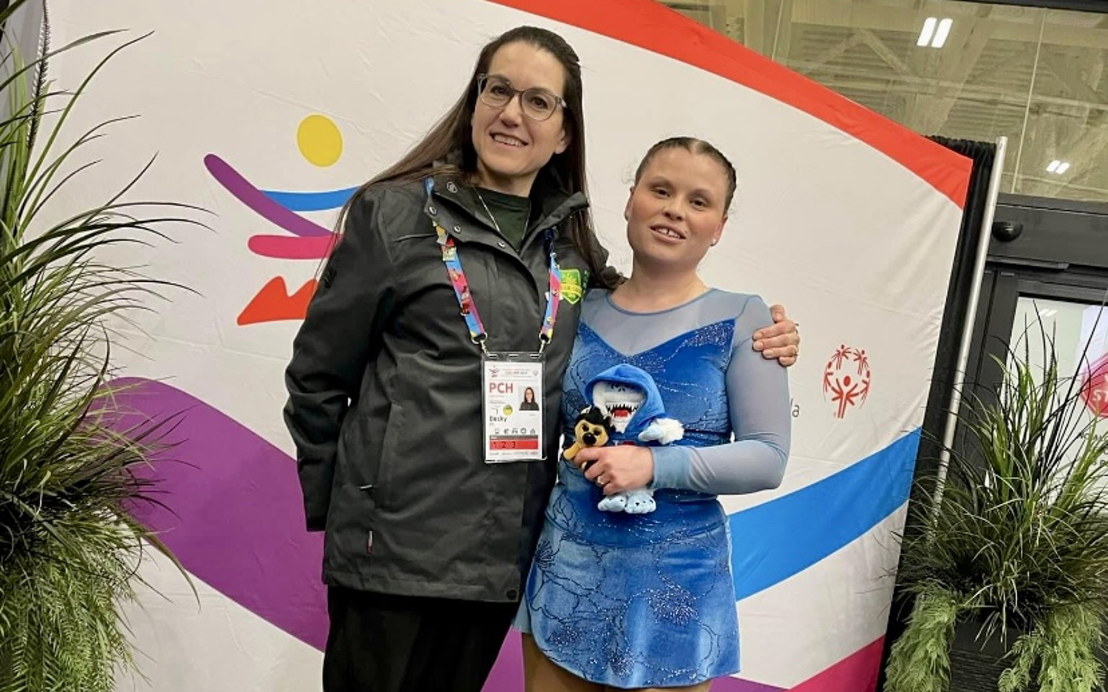 A young figure skating athlete is posing with her coach. They are both smiling for the camera and standing in front of a backdrop. The figure skater is still in her skates and blue figure skating outfit. The coach is wearing black pants and a black jacket with a colourful lanyard hanging around her neck.