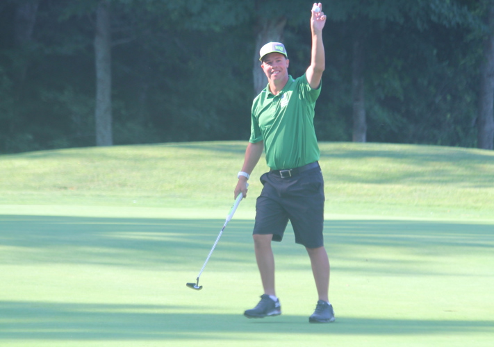 A golf athlete is standing on the golf course holding up a golf ball in one hand. In the other hand, he is holding his golf club. He is smiling for the camera and wearing a green golf shirt, grey shorts, and a grey baseball cap.