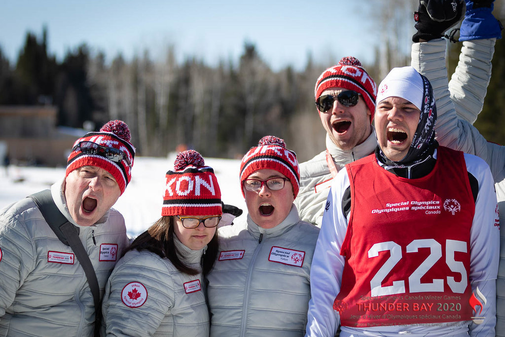 Five Special Olympics Yukon athletes are cheering for their team. They are all wearing white puffer jackets and red Yukon toques. One athlete on the far right is wearing a red pinnie with the number 225 on it.