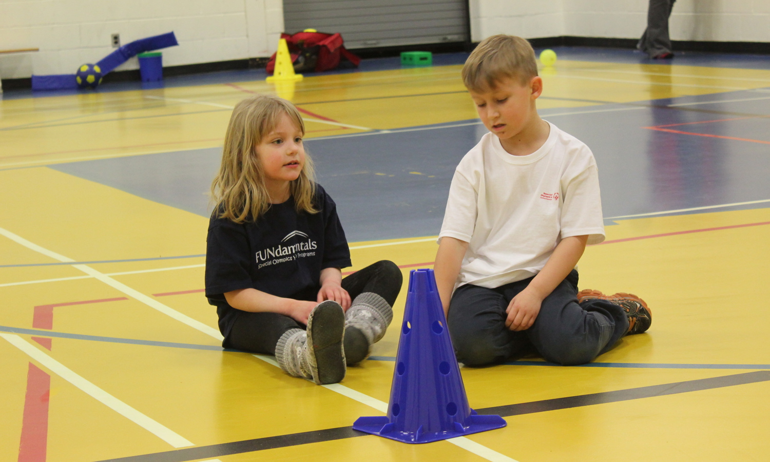Two small children are sitting on a gym floor in front of a blue pylon. The girl on the left is wearing a black FUNdamentals t-shirt and the boy on the right is wearing a white Special Olympics Saskatchewan t-shirt.