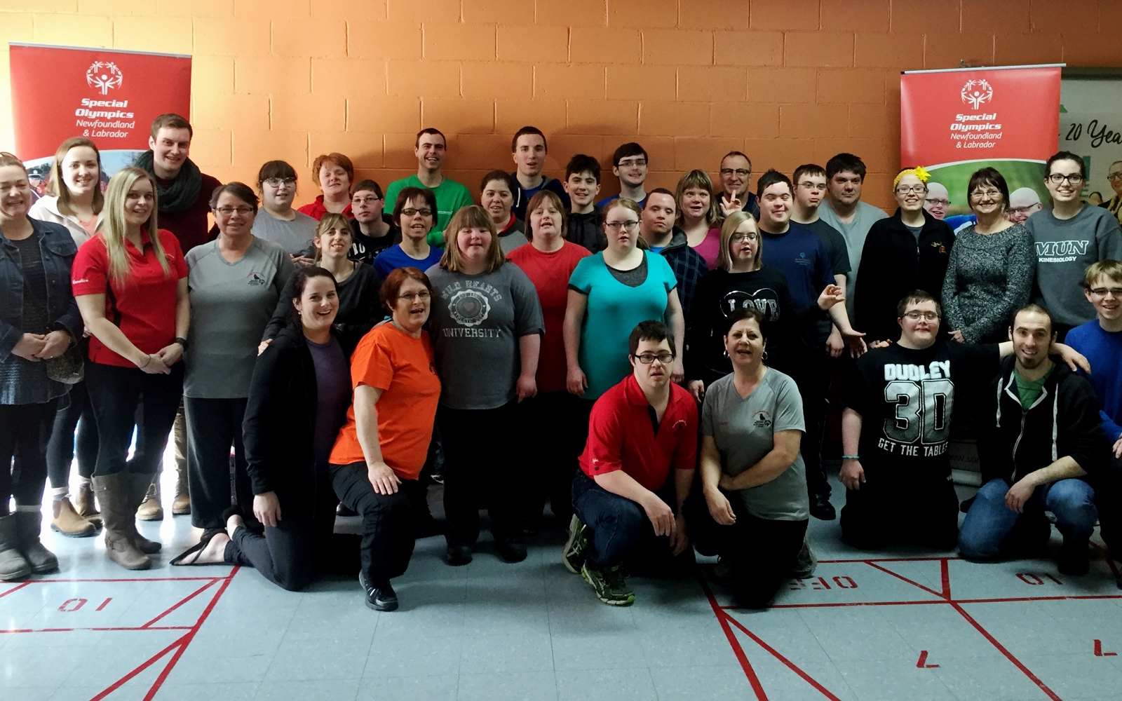 A group photo of multiple athletes, coaches, and volunteers standing against an orange brick wall. Everyone is smiling for the camera and wearing casual clothes.