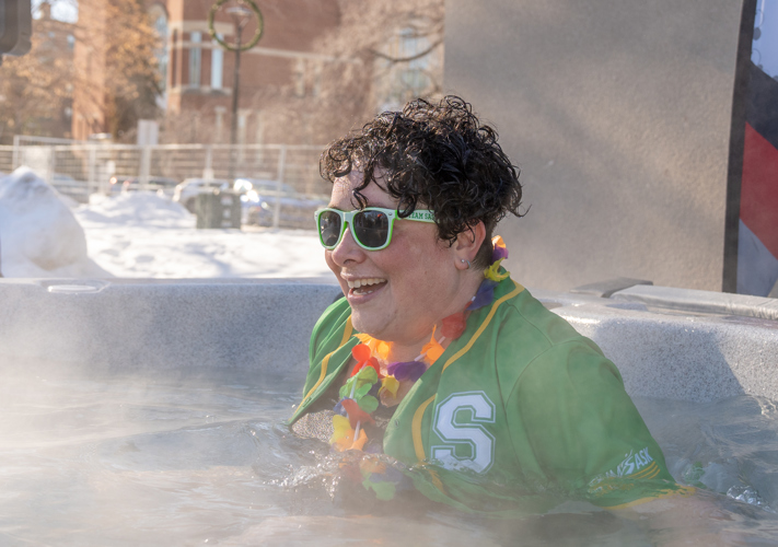 A individual taking a plunge in an ice cold water tub. She is wearing a green Saskatchewan baseball jersey and green sunglasses.