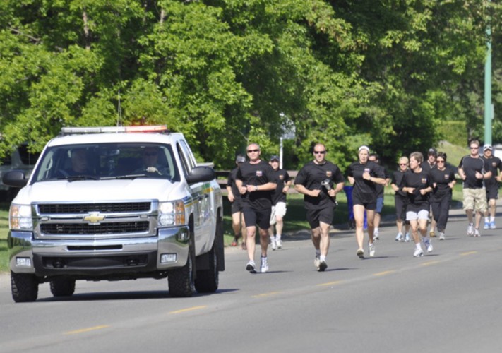 A group of Law Enforcement Torch runners can be seen running in the distance. They are all wearing black t-shirts and some are wearing sunglasses. They are running next to a white police truck with it's lights on.