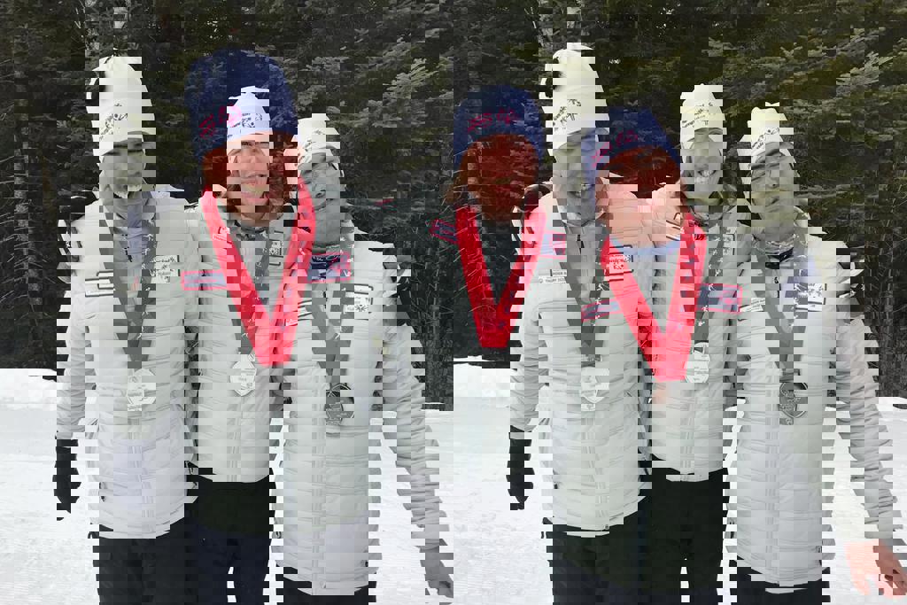 A group of three athletes hugging and smiling with silver medals around their necks. They all wear the same cream coloured jacket and white toques. There are trees in the background and snow on the floor. 