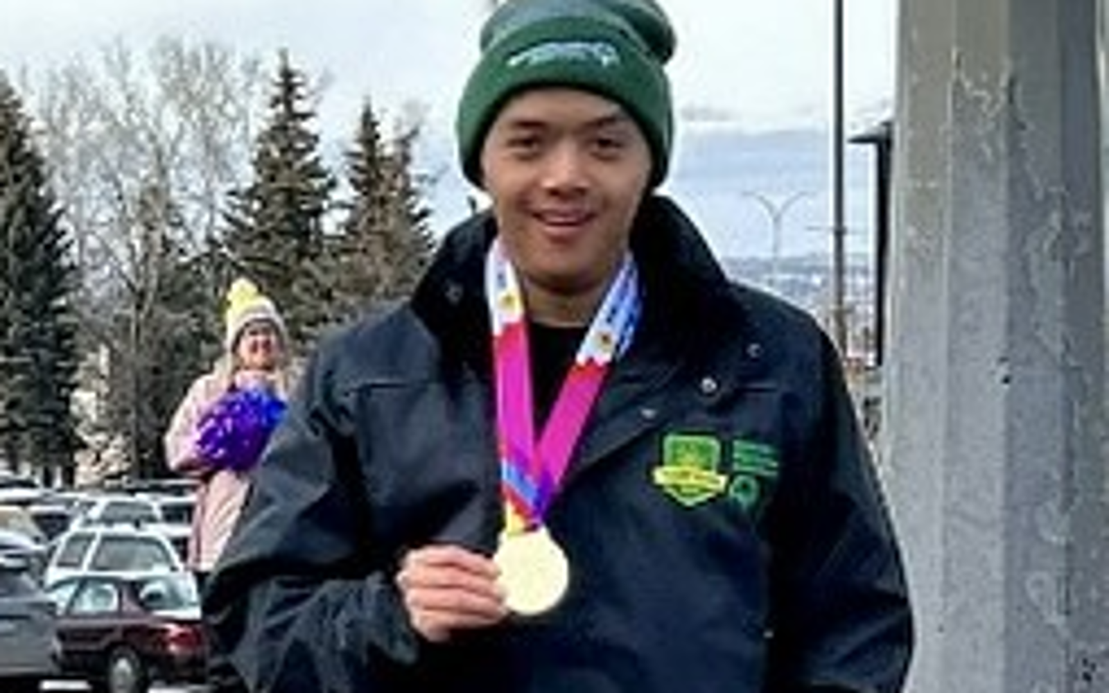 A photo of Michael, a Special Olympic Saskatchewan athlete. He is standing outside and wearing a winter jacket and toque. Around his neck is a medal and he is holding the medal with one hand. He is also smiling for the camera.