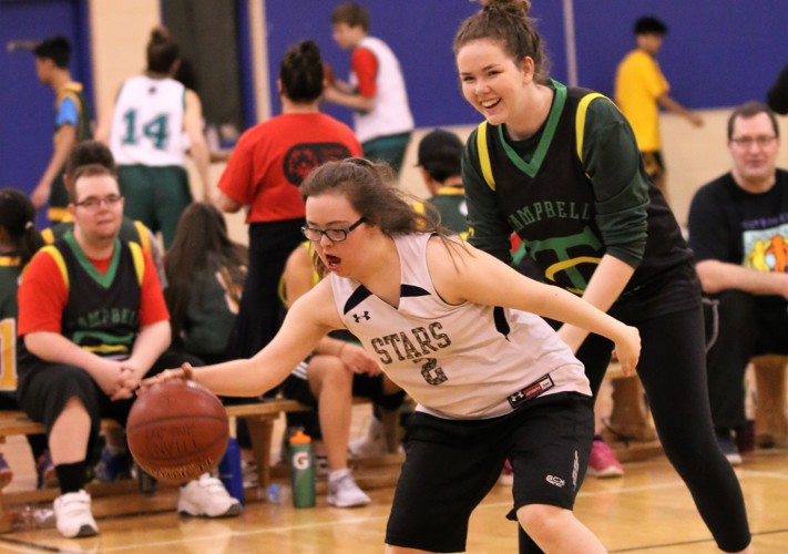 Action shot photo of two athletes playing basketball. One athlete in a white jersey has possession of the basketball. The other athlete is standing behind her and wearing a black jersey. Behind the play, you can see other athletes sitting on the bench.