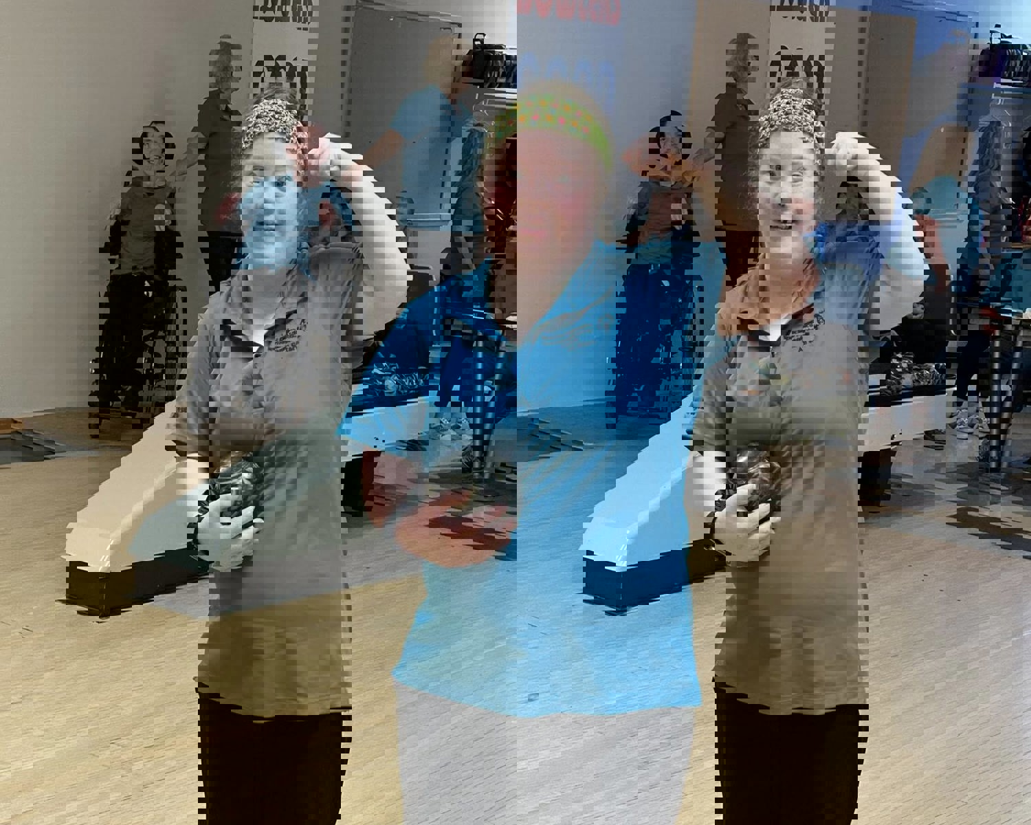 An athlete is standing at the start of the bowling aisle. She is holding a bowling ball in one arm while the other arm is raised to show off her muscles. She is wearing a blue bowling t-shirt and smiling for the camera.