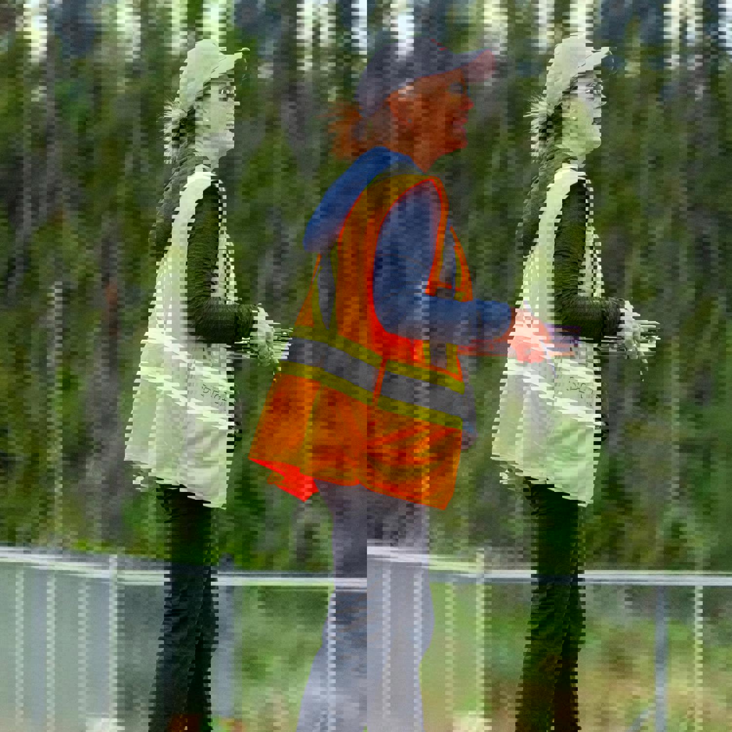 A coach, on the sideline, watching the match with her clipboard ready to take notes on what she is watching. She is wearing a high visibility vest, a baseball cap, and sunglasses.