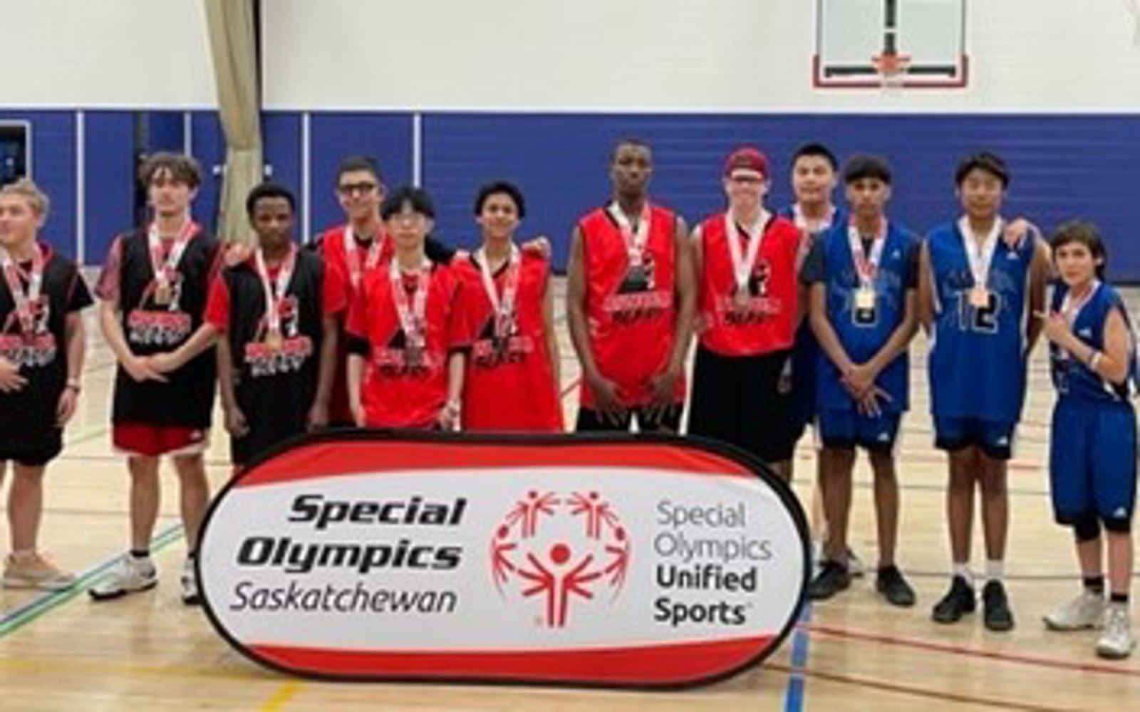 Basketball athletes posing for a group photo. They are standing in a gymnasium in front of a sign that reads, "Special Olympics Saskatchewan. Special Olympics Unified Sports." Every athlete has a medal around their necks. From left to right are athletes wearing black jerseys, red jerseys, and blue jerseys.