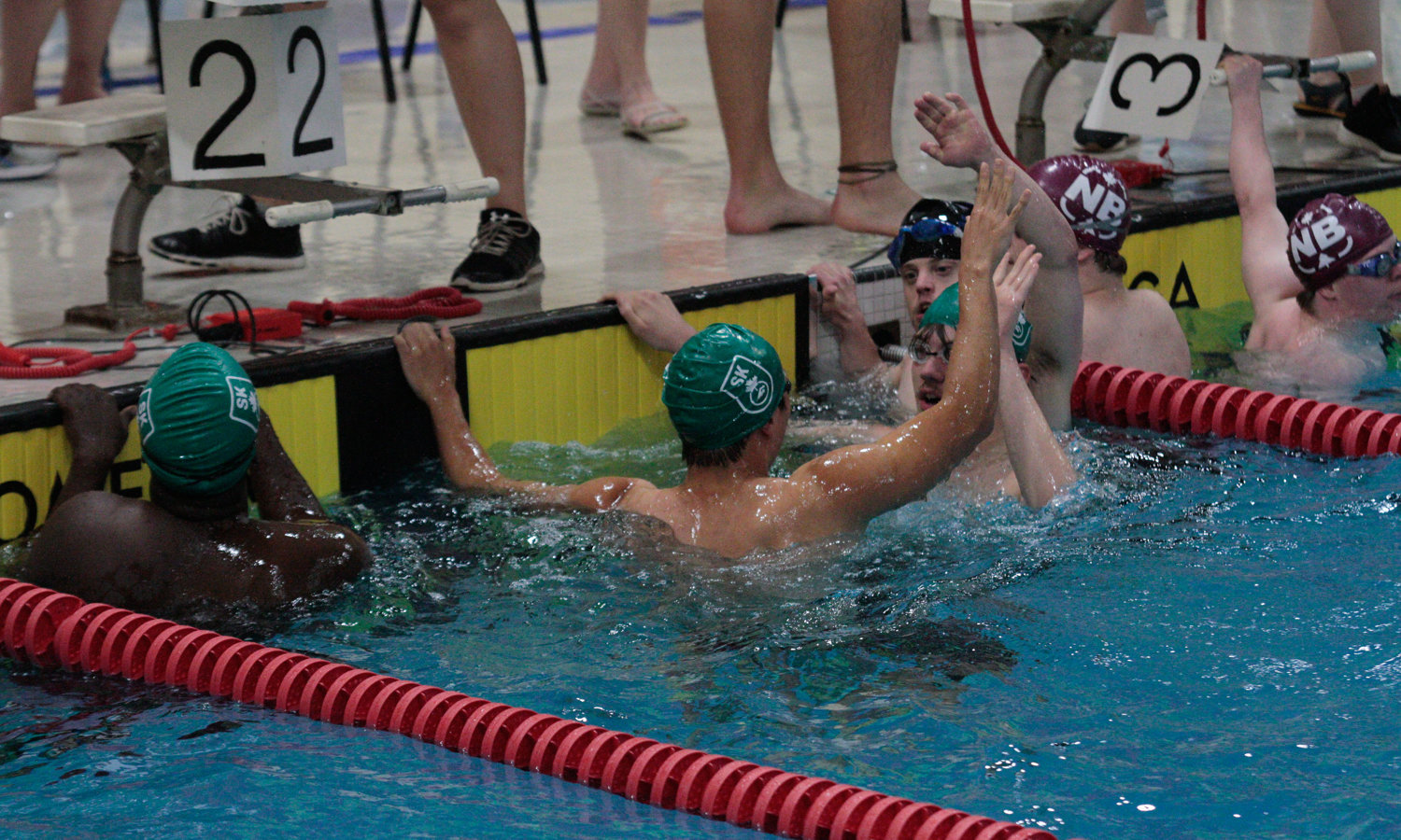 Six swimmers are holding on to the side of the pool, in the water, and giving each other high-fives. The two athletes in the front of the image have their backs turned towards the camera while you can just see the faces of the two athletes they are high-fiving.