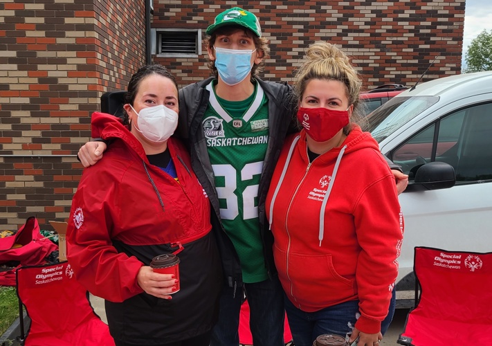 Three fundraisers preparing for an event. One individual in the middle is wearing a green Saskatchewan jersey, while the other two individuals beside him are wearing a red jackets/sweater. They are standing behind a table that has many red t-shirts on it. The three fundraisers are wearing masks.