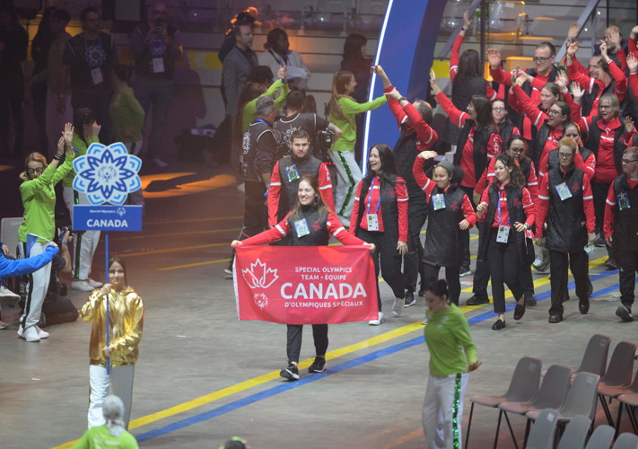 Group of athletes walking into opening ceremony. Front athlete is holding a Canadian banner with the words Special Olympics Team Canada. All athletes are dressed in red and black. 