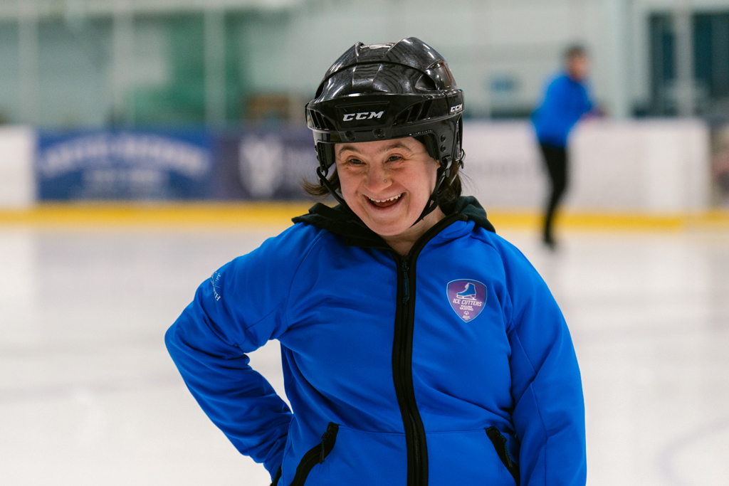 A close up image of a figure skating athlete smiling. She is on the ice and wearing a blue sweater and a black helmet.
