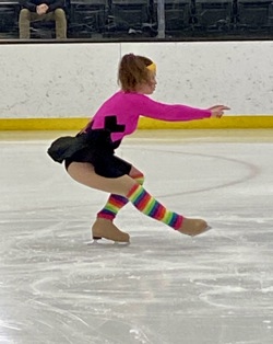 An action shot photo of a figure skater, mid routine. She is in the middle of a spin. She is the only athlete on the ice and she is wearing a pink bodysuit with a black skirt, a yellow headband, and rainbow leg warmers.