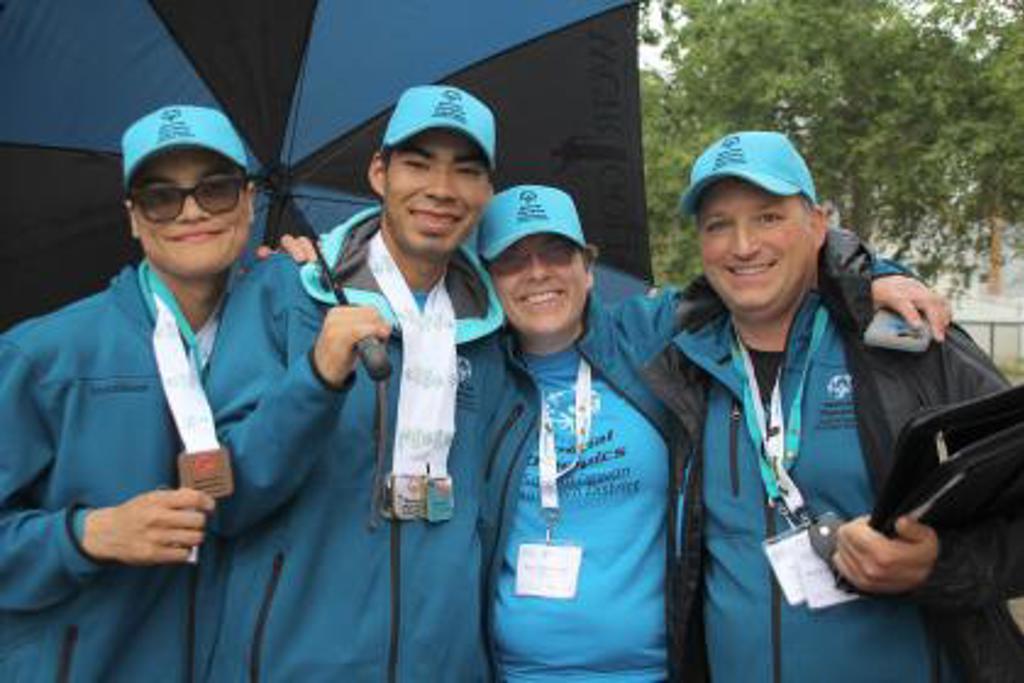 Four people, two athletes and two volunteers posing with their arms around each other. Everyone is wearing blue and has blue baseball caps on. The athletes have medals around their necks while the volunteers have a lanyard around their necks.