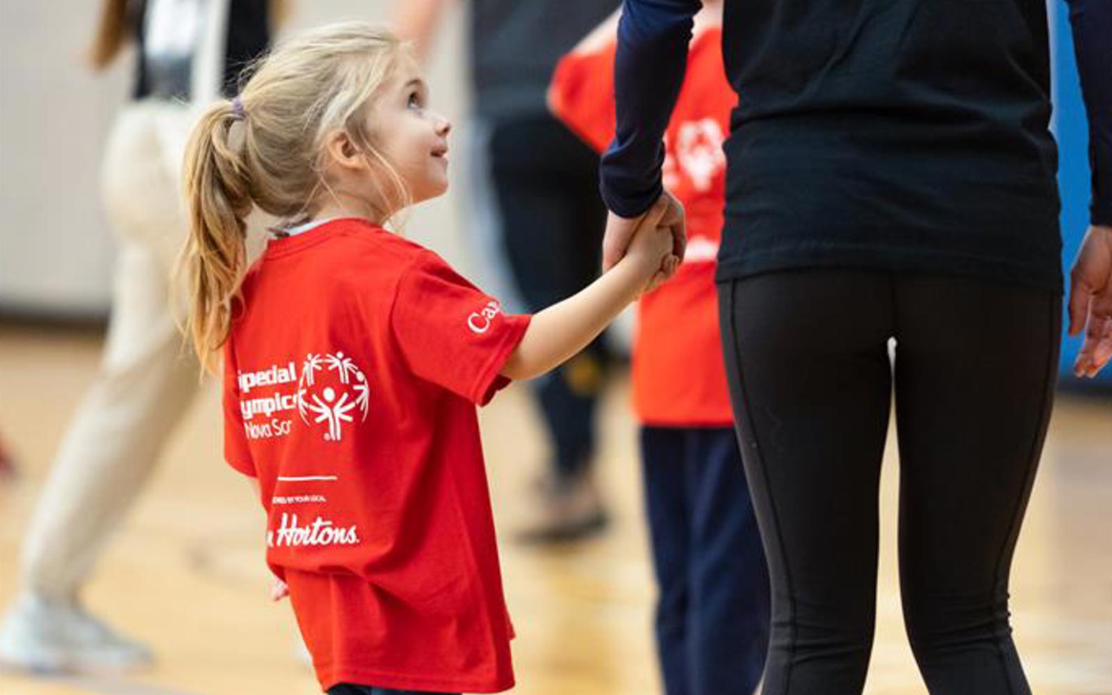 A young athlete is looking up and holding the hand of her guardian. She is wearing a "Special Olympics Nova Scotia" shirt and smiling at the individual she is holding hands with. The picture is taken from behind the pair, and they are located in a gym.