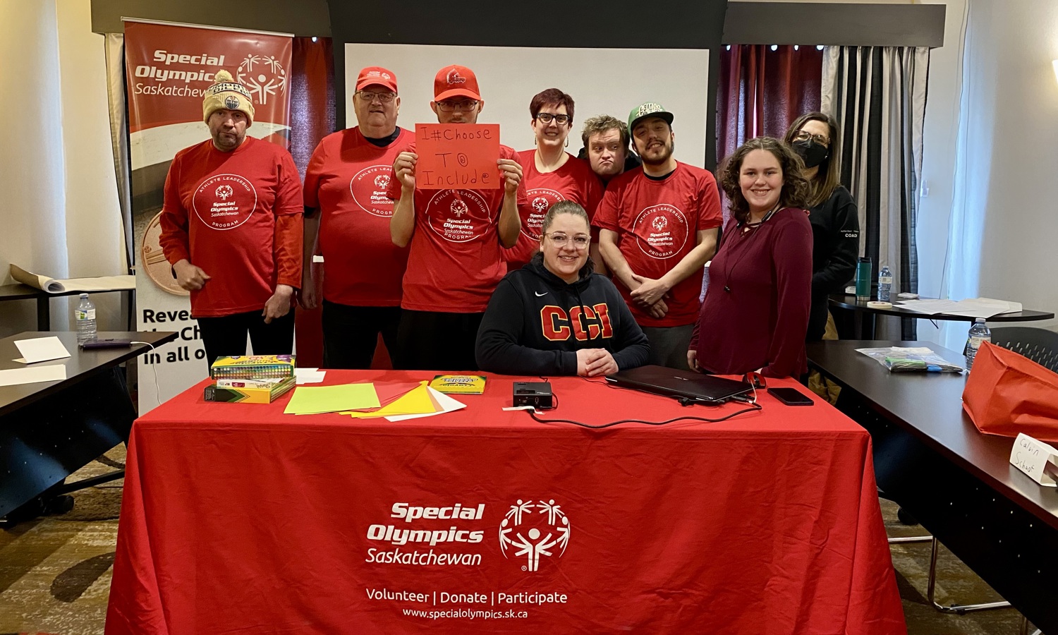 A group of athletes and volunteers are sitting and standing behind a table and smiling for the camera. Many people are wearing red Special Olympics Saskatchewan long sleeved shirts.