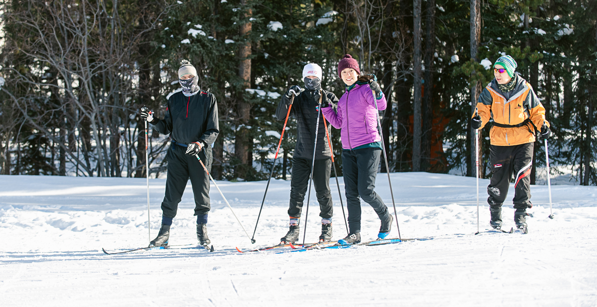 Four cross country skiers are smiling for the camera. They are wearing winter attire, jackets and hats, and have their skis on.