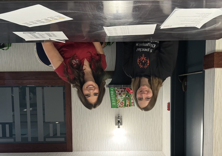 Two volunteers are sitting behind a table and smiling for the camera. In front of them are stacks of paper.