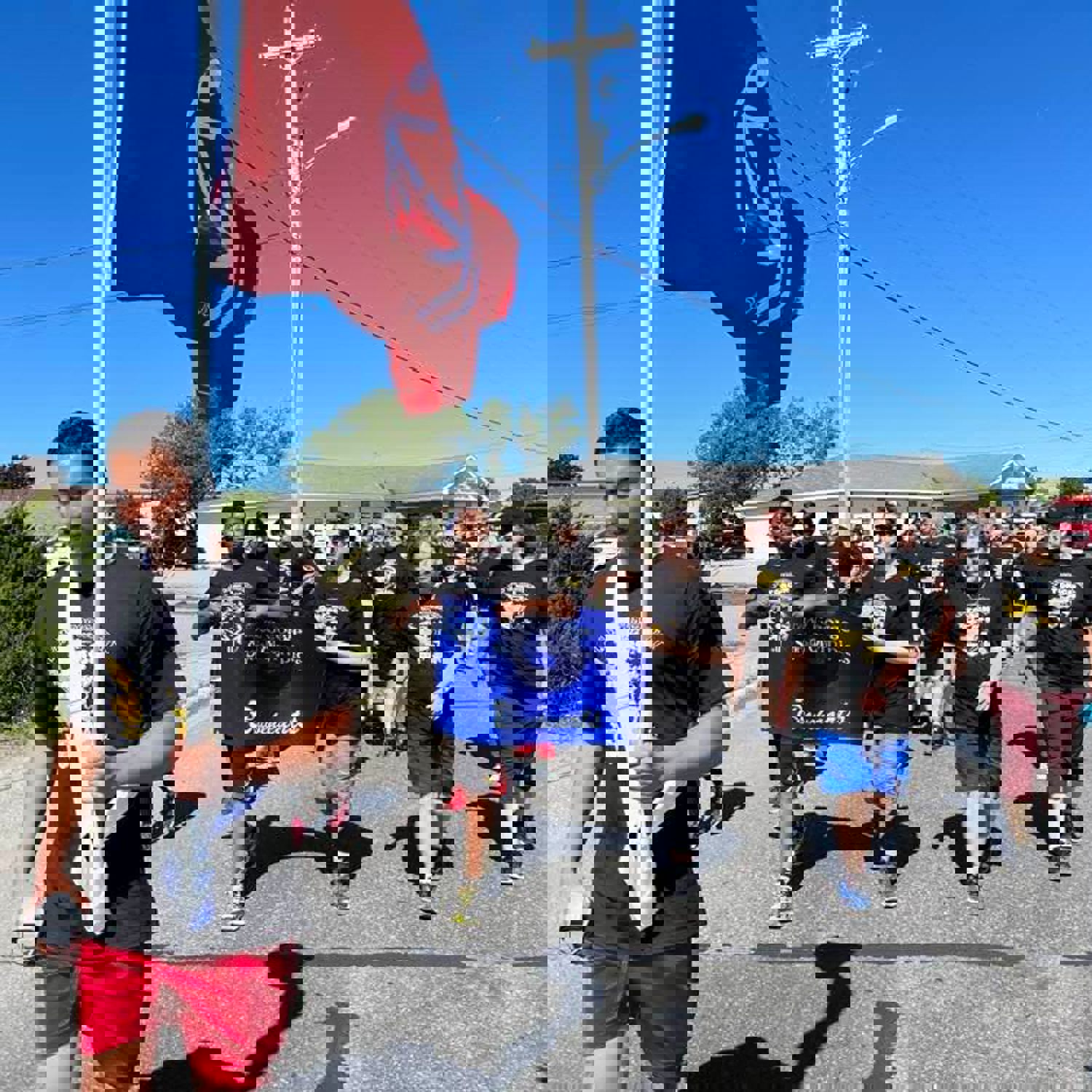 Law Enforcement Torch Runners are walking down the street in matching Law Enforcement Torch Run t-shirts. The leader of the group is holding up a large, red, Special Olympics flag. The people behind him are holding a "Bay St. George Special Olympics Bravehearts" banner.