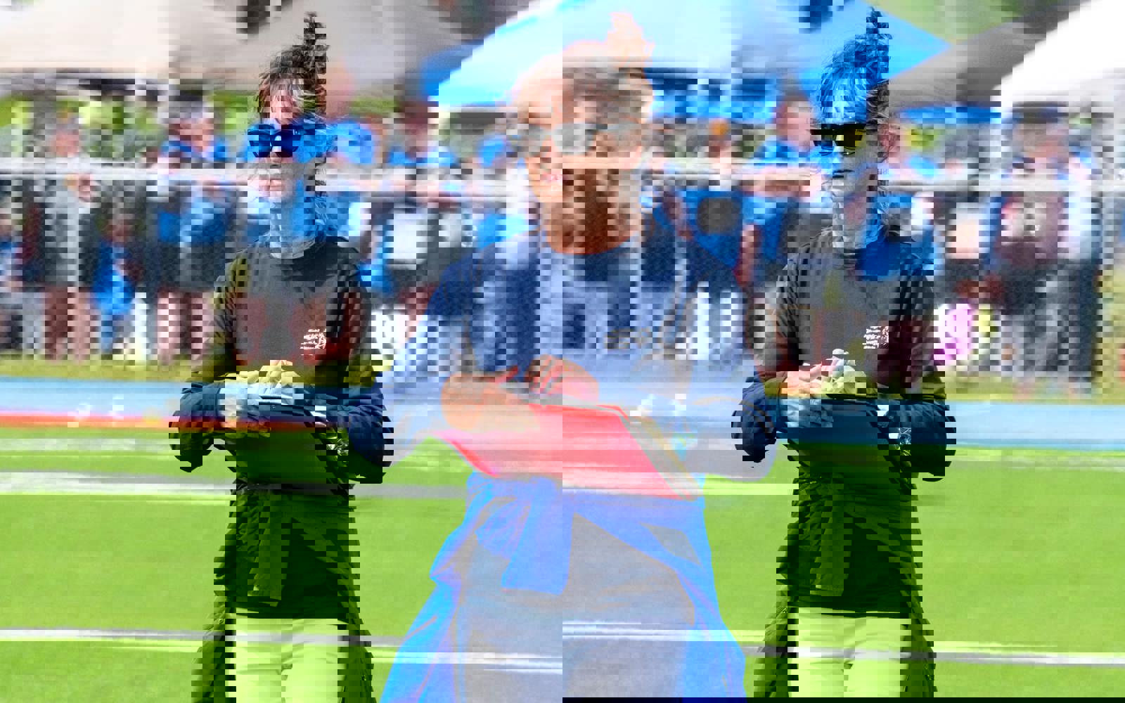 A volunteer with her red binder, is standing in the middle of the field. She is wearing sunglasses and a navy blue long sleeve shirt. She is wearing a light blue jacket tied around her waist.