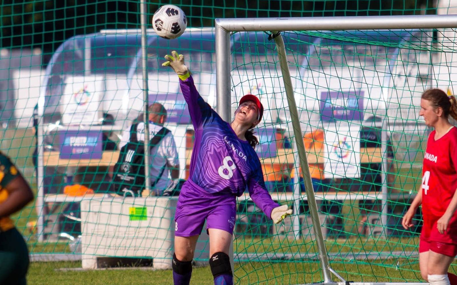A soccer goalie standing in front of her soccer net, throwing the ball out onto the field. She is wearing a purple long sleeved jersey with Canada and the number 8 written on the front of it.