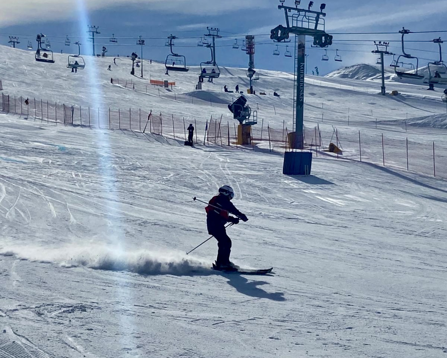 An action shot photo of an alpine skier going down the hill. The skier is a medium distance away from the camera and you can see the snow blowing behind their skis. In the distance behind the skier, you can see the ski lift and other skiers going down a different hill.