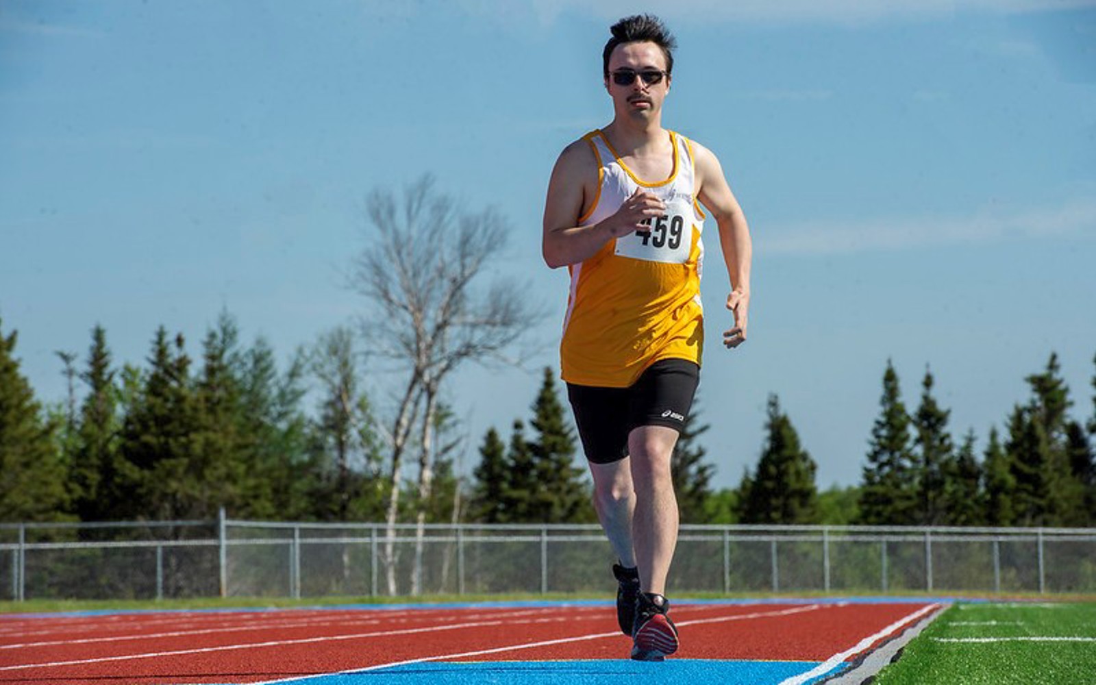An athletics athlete is running down a track. He is the only athlete on the track. He is wearing a yellow tank top, with the number 459 pinned to it, as well as sunglasses, and black athletic shorts.
