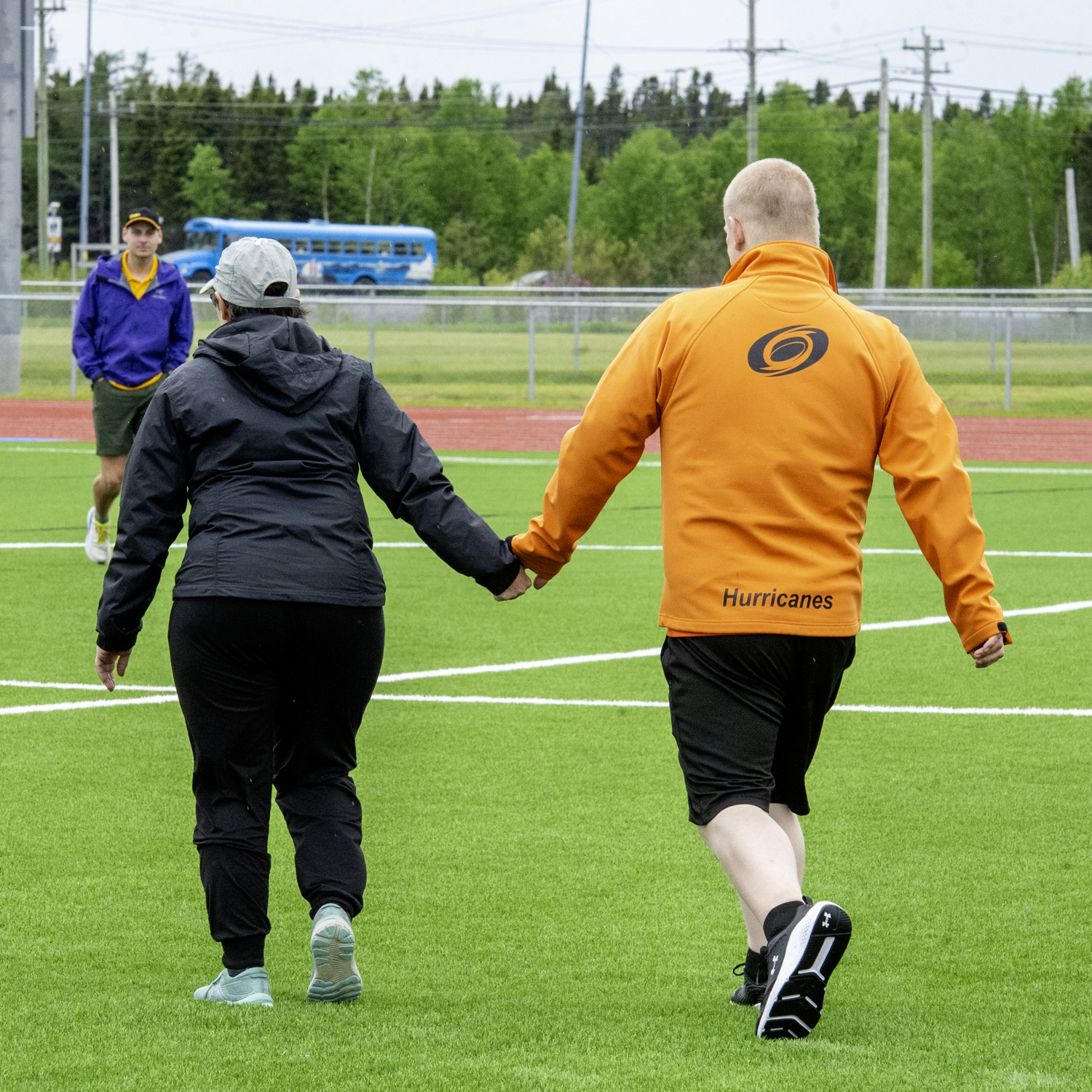 Two athletes holding hands walking on the field. The photo was captured from behind the athletes so you cannot see their faces. They are both wearing light rain jackets to prepare for the weather.