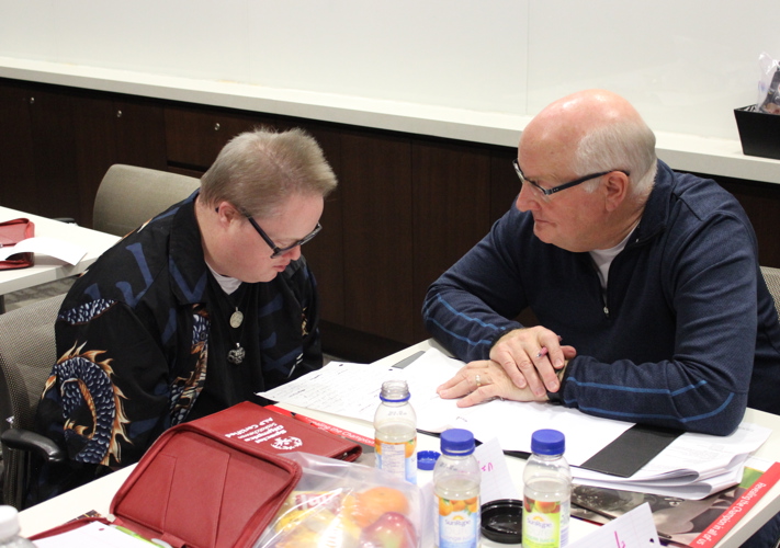 Two individuals are sitting at a table covered with papers and snacks. The individual on the right is reading something off the paper to the individual on the left.