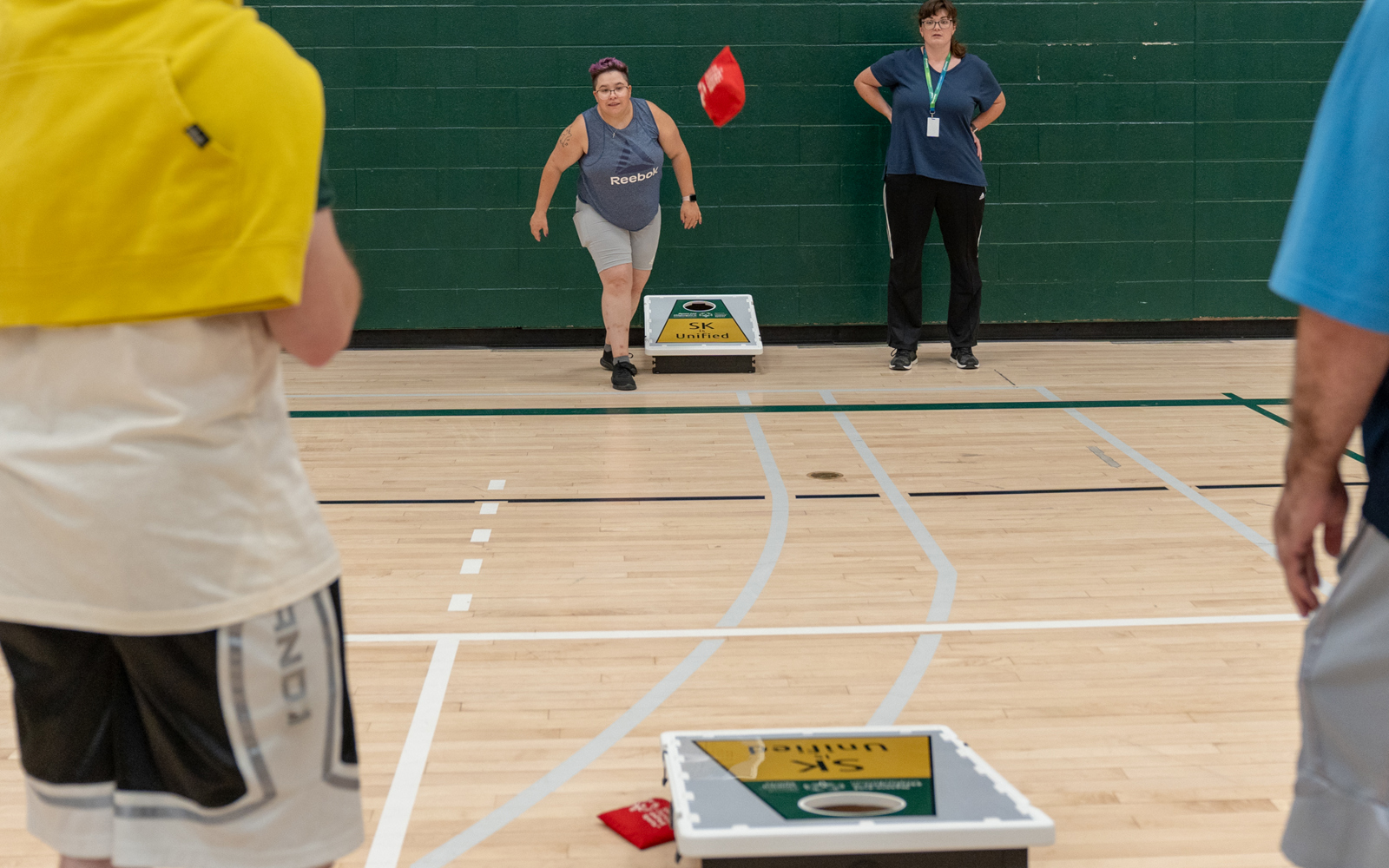 An action shot photo of people playing bean bag toss. In the distance you can see someone throwing a bean bag. The bean bag can be seen in midair coming closer to the camera. On the floor are the bean bag boards where the athletes are trying to toss the bean bags into. The game is being played inside a gymnasium.