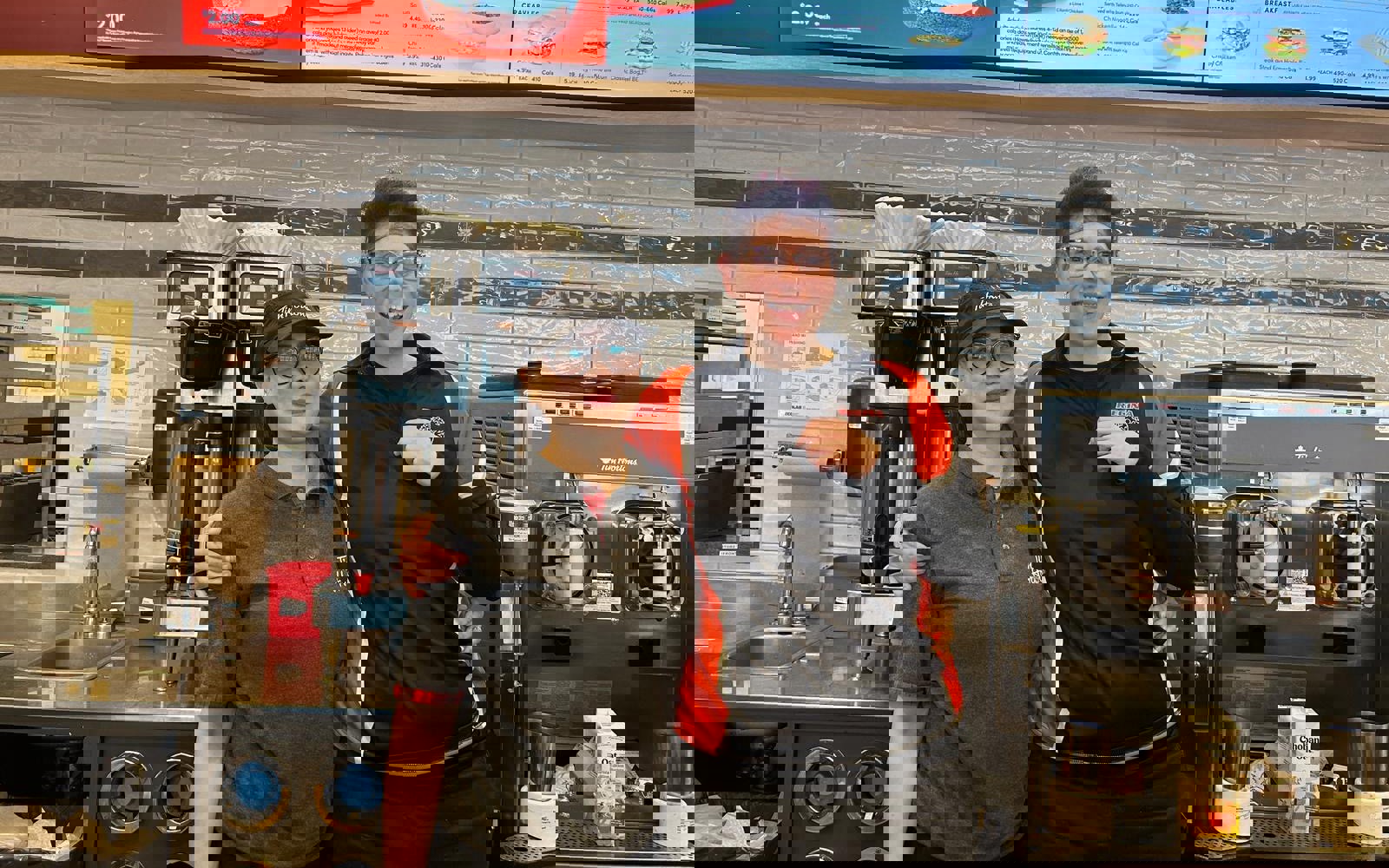 A female Special Olympics Saskatchewan athlete poses for the camera with a Tim Hortons employee. They are behind the Tim Hortons counter with their arms wrapped behind each other and they are smiling. Behind them are the display screens as well as coffee makers and equipment.