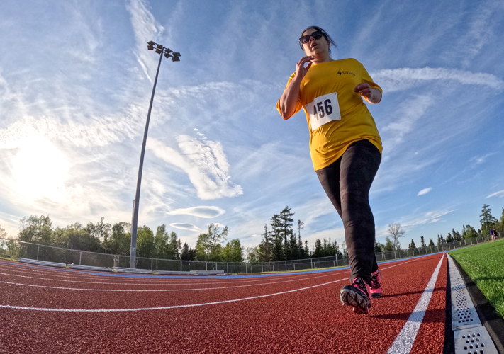 A slightly warped, low angled action shot photo of an athletics athlete running on the track. It is a bright, partly cloudy day and the athlete is wearing a yellow t-shirt with the number "456" pinned to the front of it. She is also wearing sunglasses and black and pink running cleats.