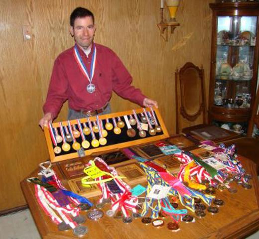 Athlete Tyler Repka's showing all of his athletic achievements. He is wearing a red button down shirt, with a medal around his neck. He is holding up a board lined up with multiple medals. He is also standing behind a table that has additional piles of plaques and medals he has received. 