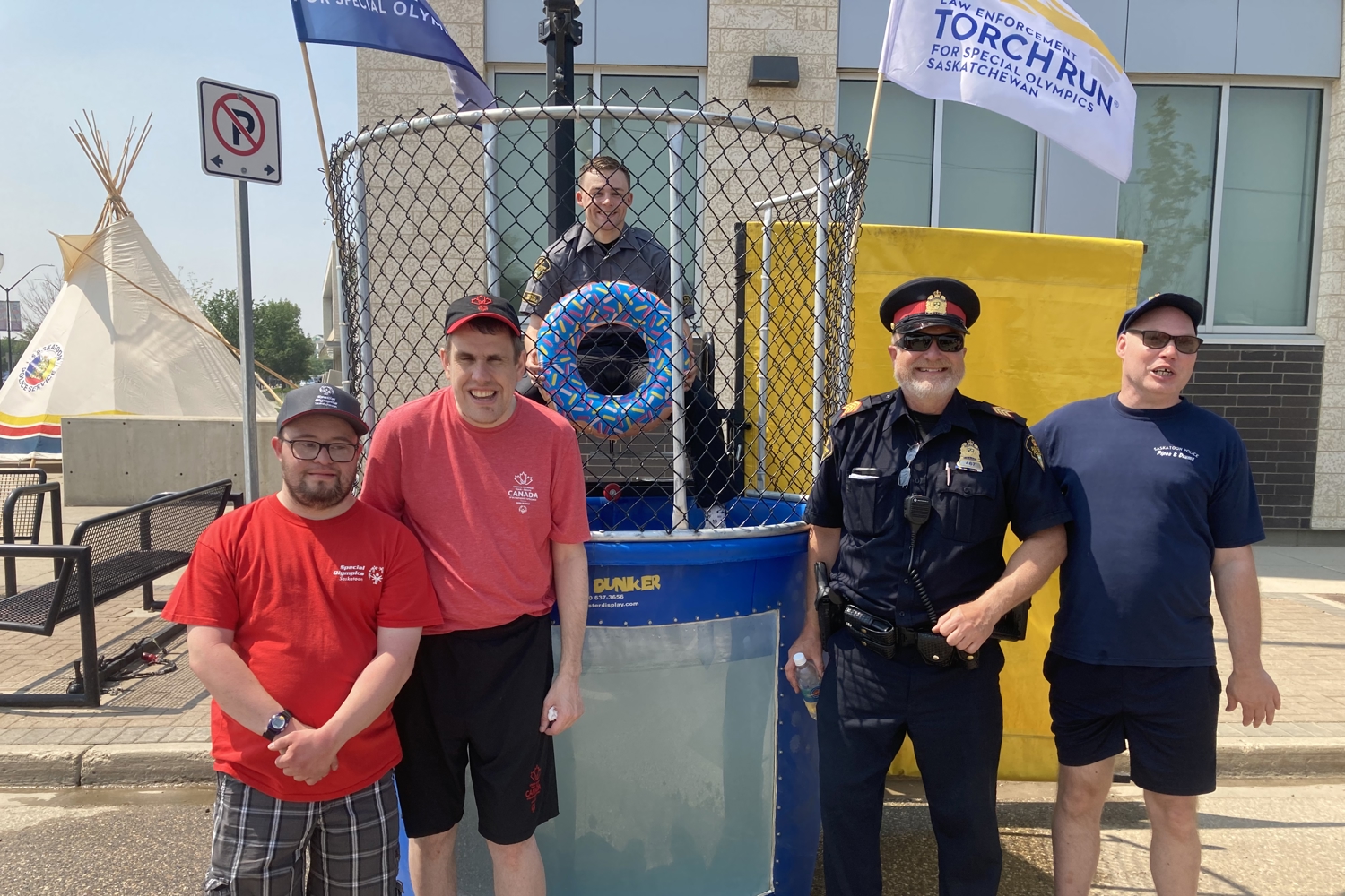 Three Special Olympics athletes are smiling and posing for the camera alongside two police officers. They are standing in front of a dunk tank and one of the police officers is inside the dunk tank.