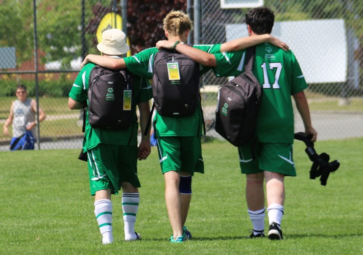 Three Special Olympics Saskatchewan soccer players, are walking off the field with their arms on each others shoulders. They are walking away from the camera so we can only see their backs. They are wearing their all green soccer uniform and have black backpacks on.