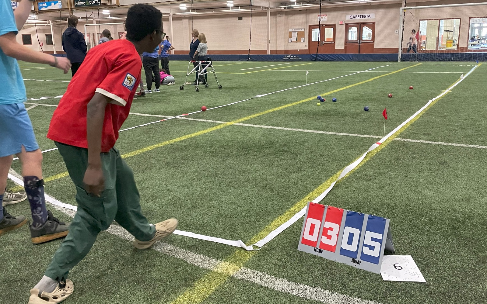 An action shot photo of an athlete playing bocce. He is watching as the ball he threw lands on the grass in front of him. You can only see the athlete from behind. On the field, you can see in the distance the other bocce balls that are in place. Next to the space defined for the game, is a scoreboard reading "03" for the red team and "05" for the blue team.