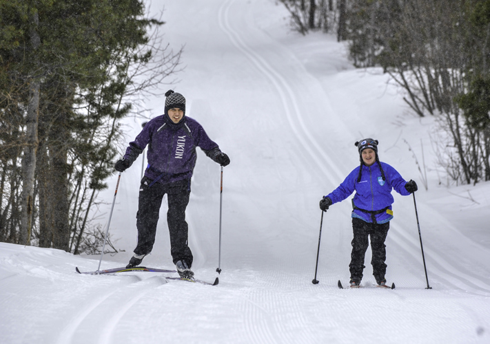 Two athletes are skiing up a hill. There is snow on the ground and trees lining both sides of the image. The athlete on the left is wearing a purple jacket that says "Yukon" on it. The athlete on the right is wearing a bright blue jacket.